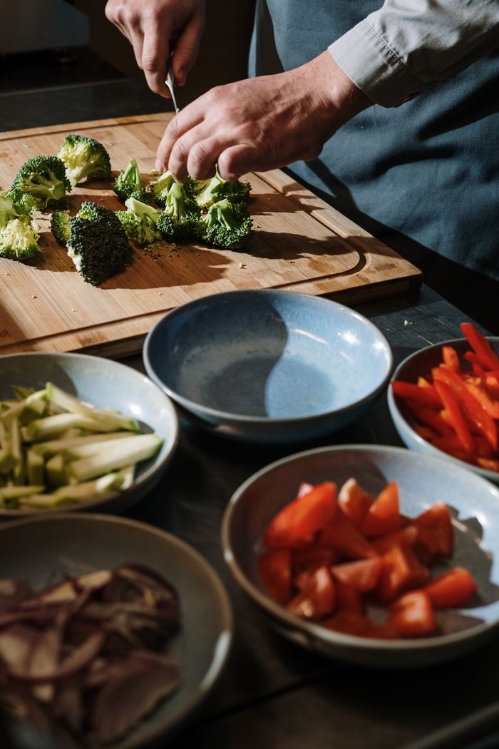 why-choose-us A chef chops broccoli on a cutting board surrounded by fresh vegetables in a kitchen setting.