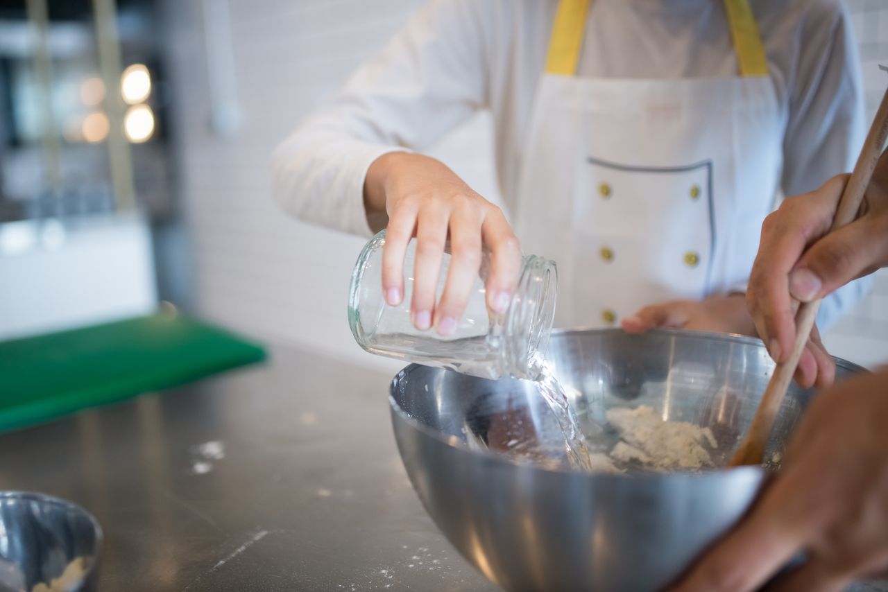 A child pours water into a stainless steel bowl, assisting in baking preparation.